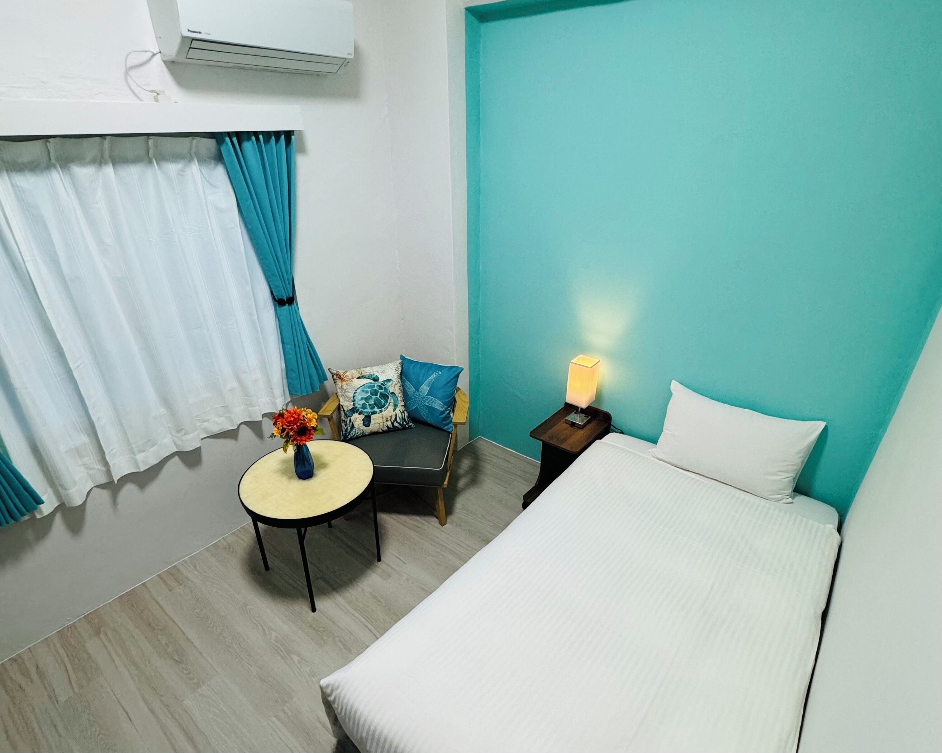 Habitación de hotel con cama, una mesita redonda con un florero, una silla con cojines, una mesita de noche con lámpara y una ventana con cortinas.