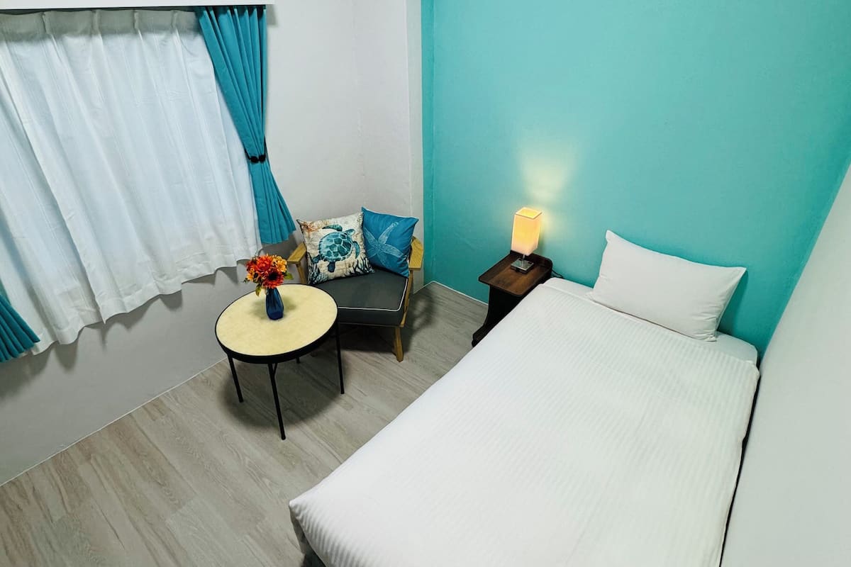 Habitación de hotel con cama, una mesita redonda con un florero, una silla con cojines, una mesita de noche con lámpara y una ventana con cortinas.