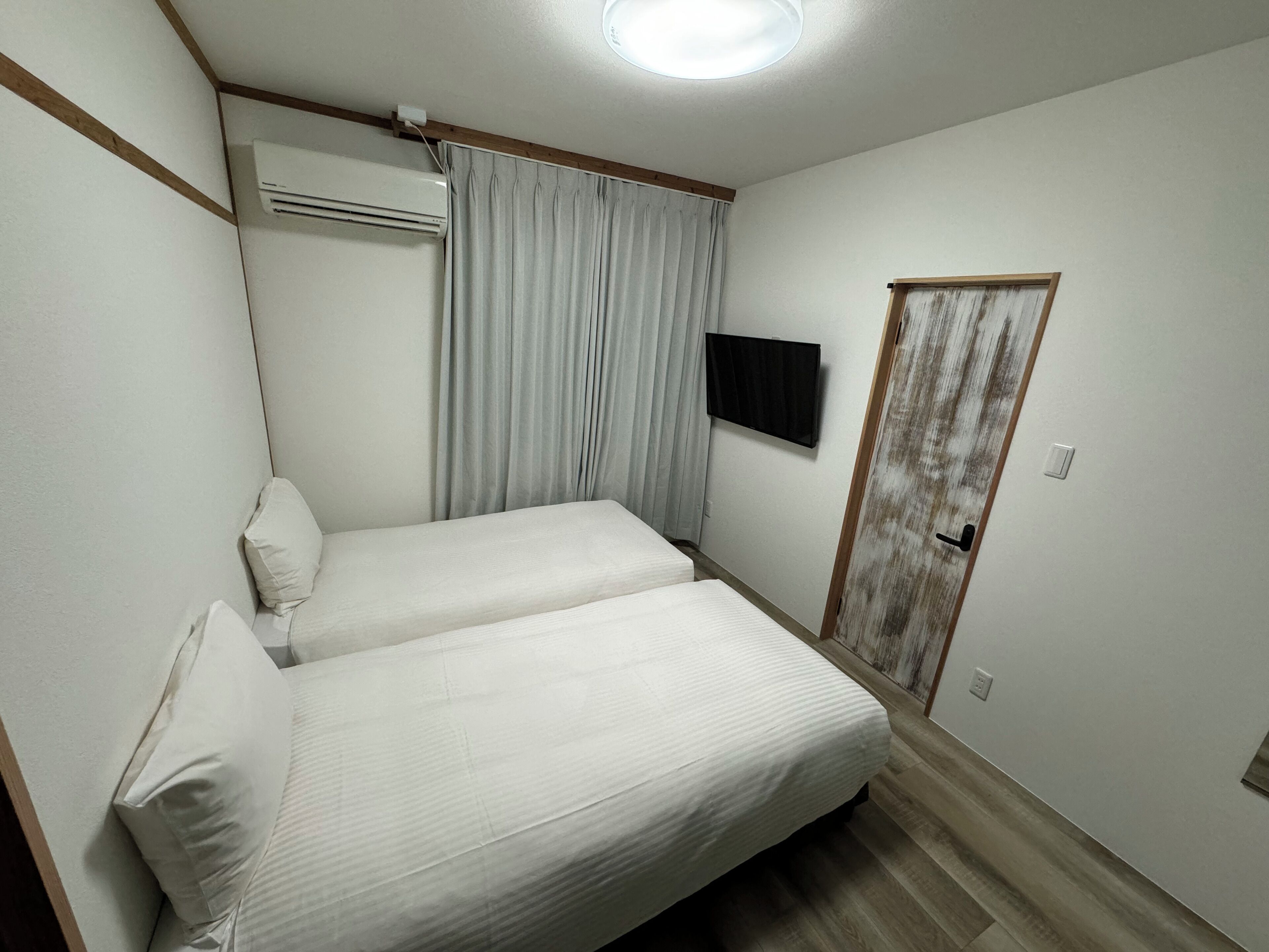 A small, clean room with two single beds, a flat-screen TV mounted on the wall, and a door with a rustic finish.