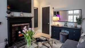 A living room with a fireplace, a flat-screen TV mounted above it, a glass coffee table with a vase of flowers, a dark-colored sofa, and a side table with a lamp. - Cosy King Suite close to Manchester City Centre, Room 2 (Stockport)