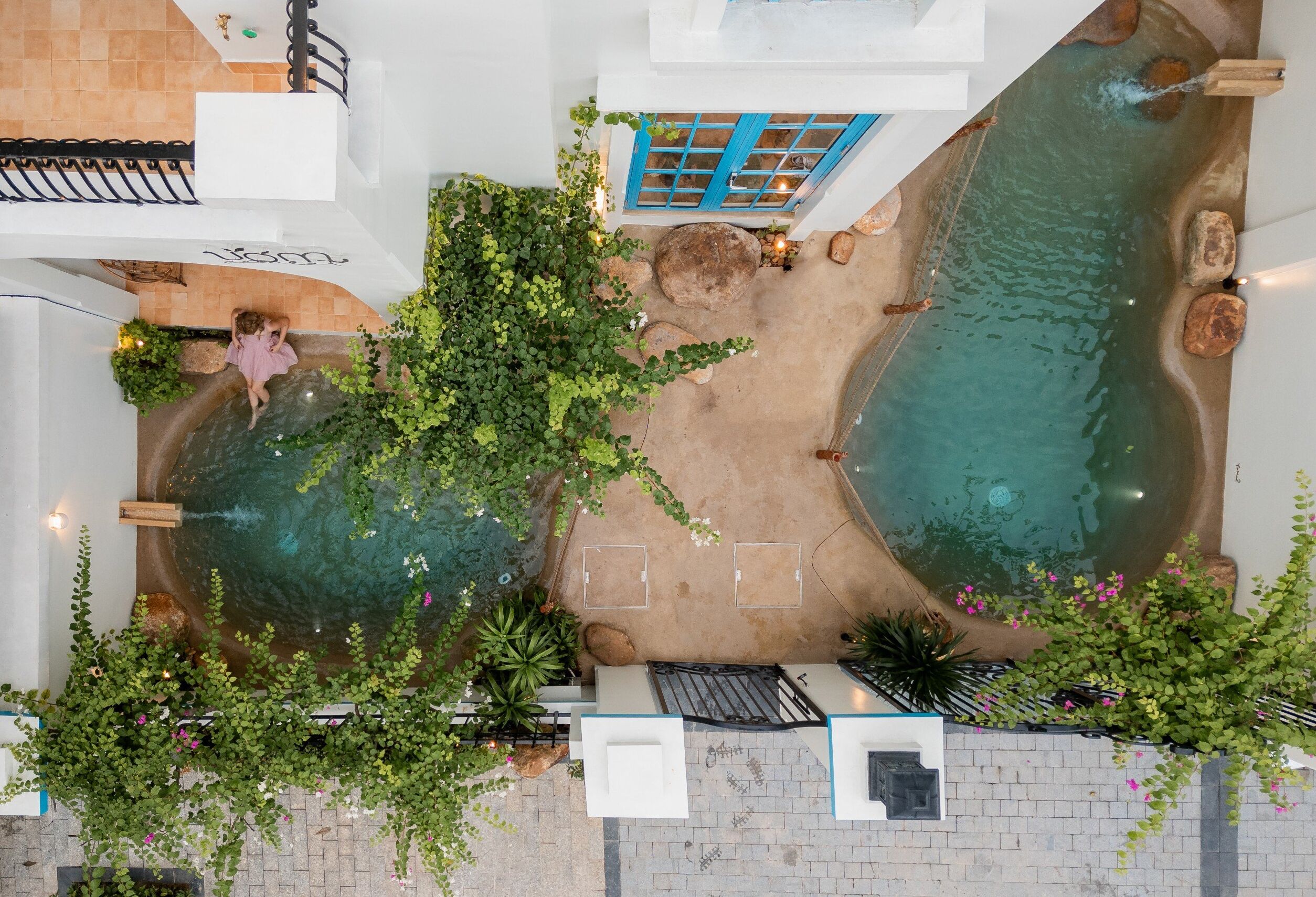 Vista aèria d'un pati amb dues piscines, envoltat de vegetació i un edifici blanc amb una porta blava.