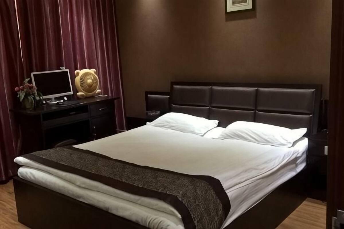 A hotel room with a large bed, a desk with a computer, a chair, and a small table with a lamp.