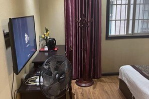 A hotel room with a bed, a TV, a fan, a window with bars, and a curtain.