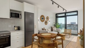 A modern kitchen with white cabinetry, a built-in microwave, and a stainless steel refrigerator. There is a dining area with a round wooden table and wicker chairs. The room has a large window with a view of the city.