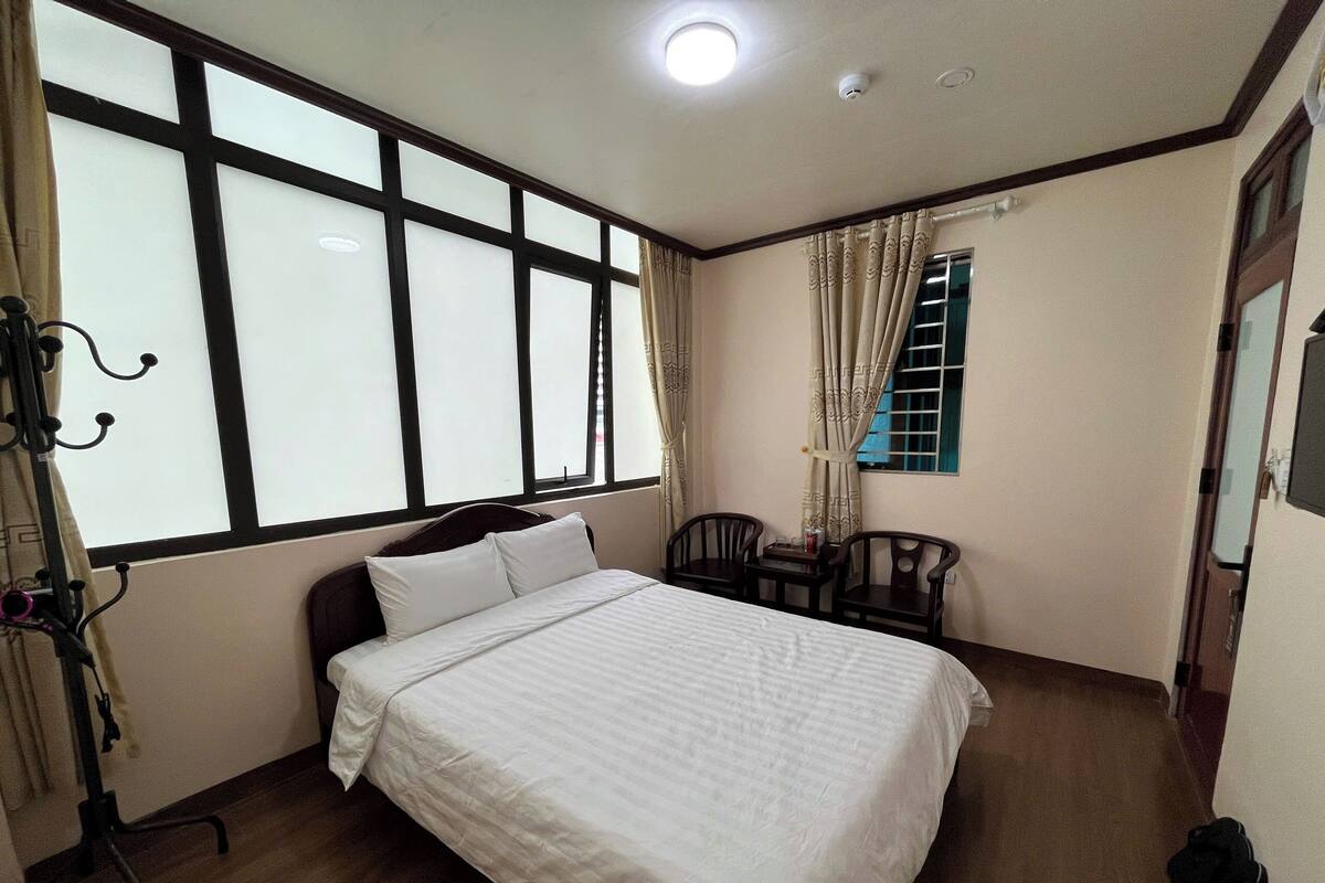 Un dormitorio con cama, una silla, una mesita, una ventana con cortinas y una puerta.