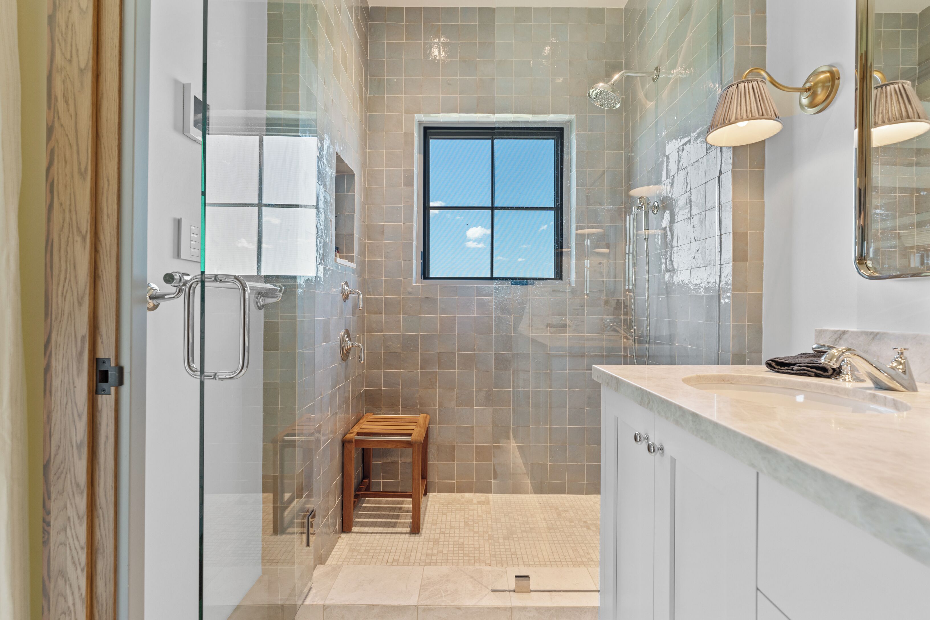 Une salle de bain moderne avec une douche à parois de verre, un petit tabouret en bois, un meuble-lavabo et une fenêtre.