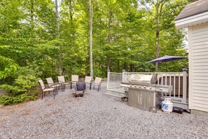 Un espace patio équipé d’un barbecue, de meubles pour se détendre en plein air et d’une terrasse offrant une vue sur une zone boisée.
