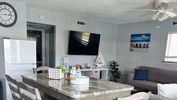 Una cocina moderna con encimera de mármol, sillas de madera, refrigerador empotrado, televisor de pantalla plana y ventilador de techo.