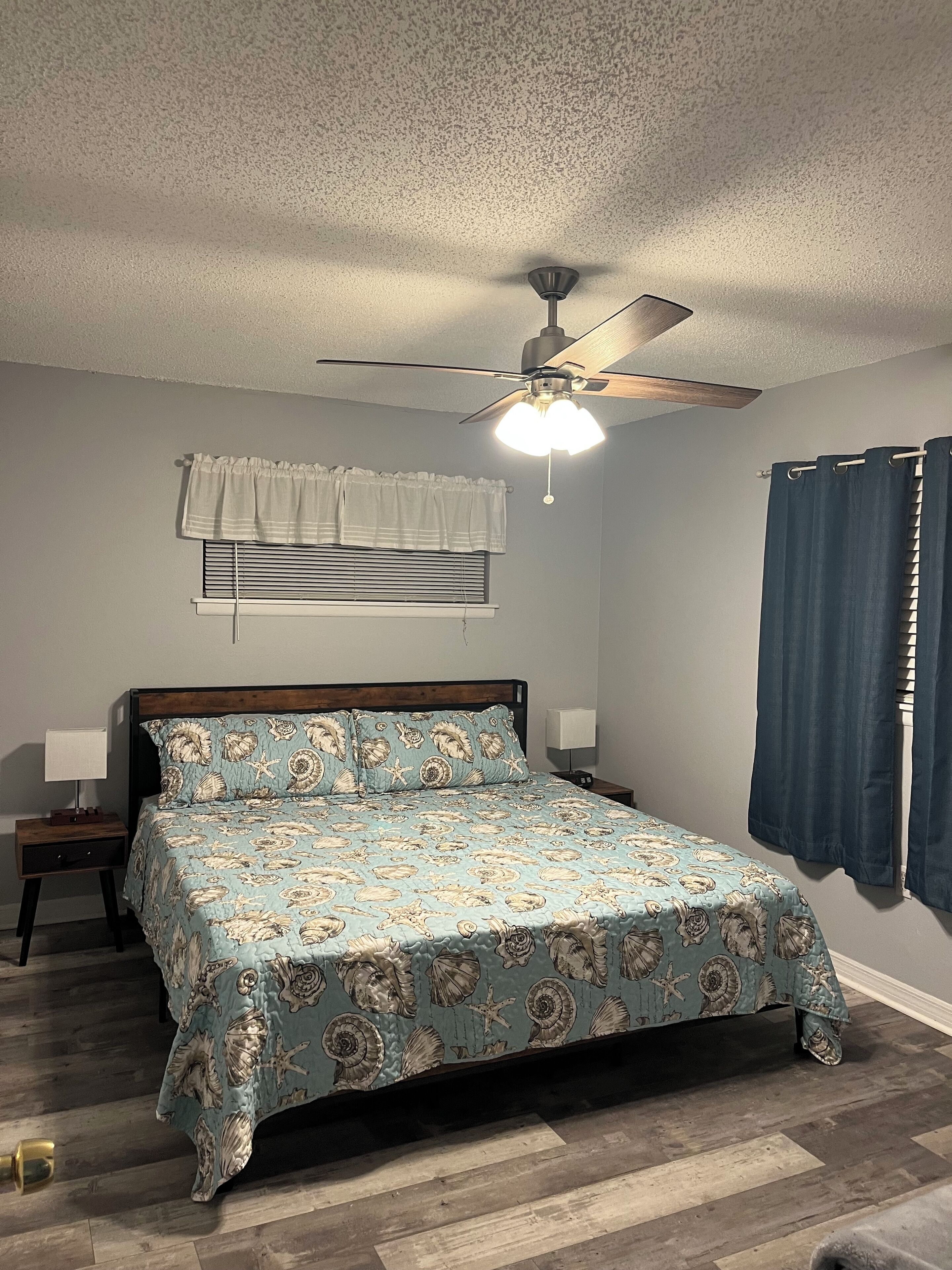 A bedroom with a bed, a ceiling fan, a window with blinds, a nightstand, and a wall-mounted lamp.