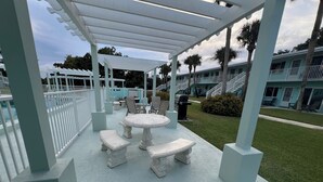 Un patio techado al aire libre con una mesa redonda y bancos, rodeado por una cerca blanca.