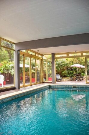 Una piscina interior con paredes de vidrio transparente, rodeada de mobiliario exterior y vegetación.
