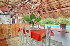 An outdoor dining area with a thatched roof, a pool, and a wooden bar.