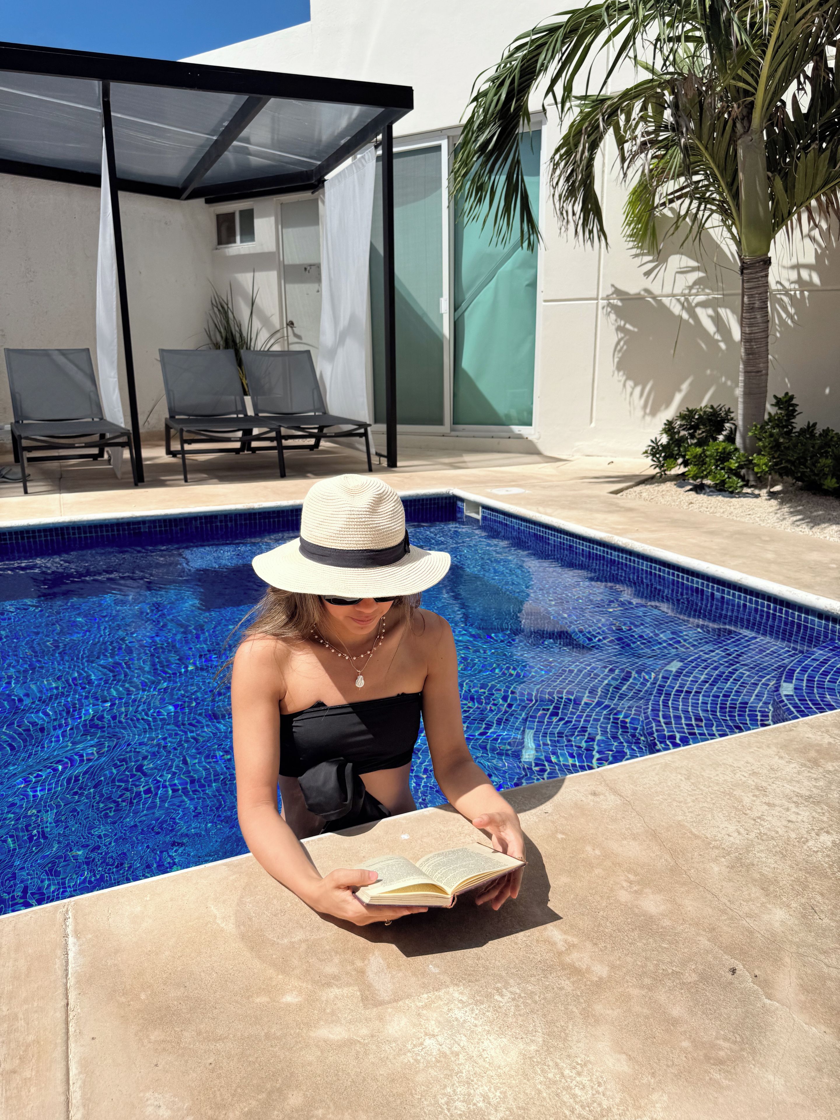 A person reading a book by a swimming pool with lounge chairs and a palm tree in the background.