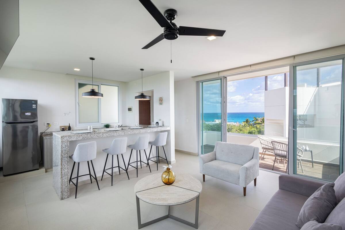 A modern kitchen with a bar, a dining area with chairs, a sofa, and a view of the ocean.