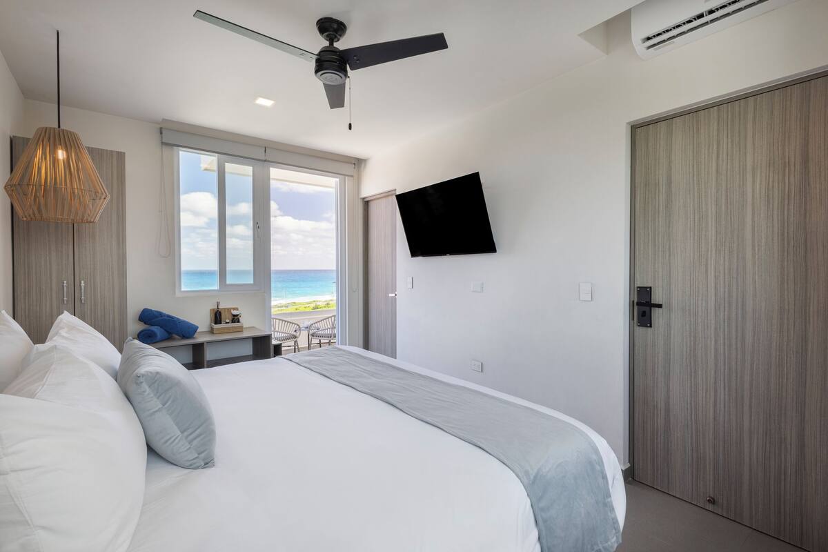 A modern bedroom with a large bed, a ceiling fan, a TV mounted on the wall, and a view of the beach through the window.