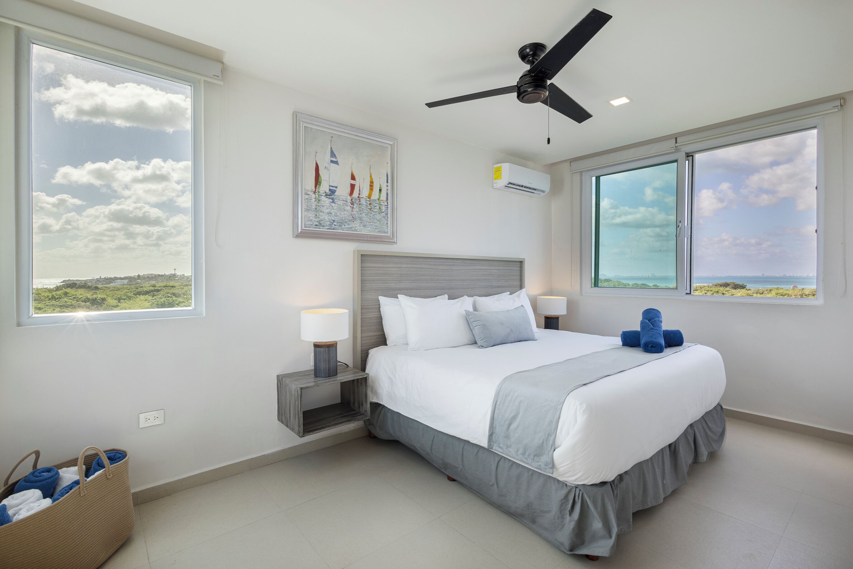 A modern bedroom with a large bed, two bedside tables, a ceiling fan, and a painting of sailboats.