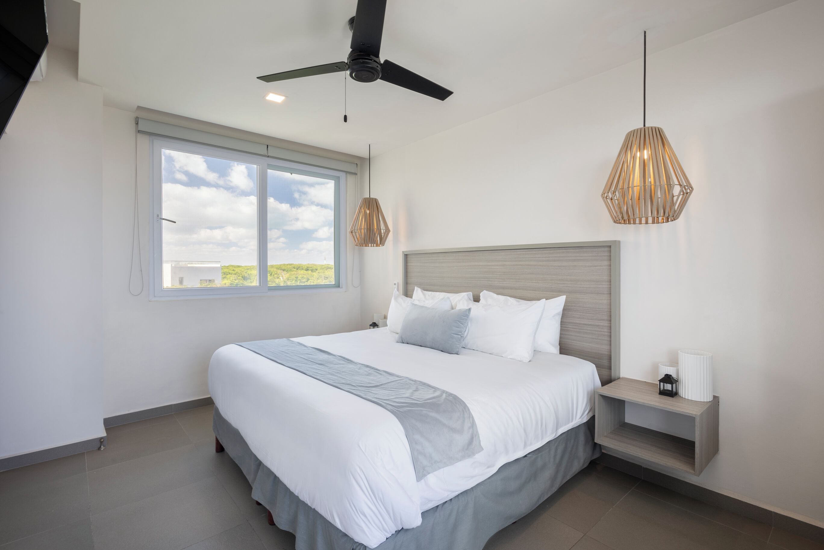 A modern bedroom with a large bed, a ceiling fan, a window with a view, and two pendant lights.