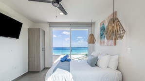 A modern hotel room with a large bed, a ceiling fan, a TV, and a view of the ocean.