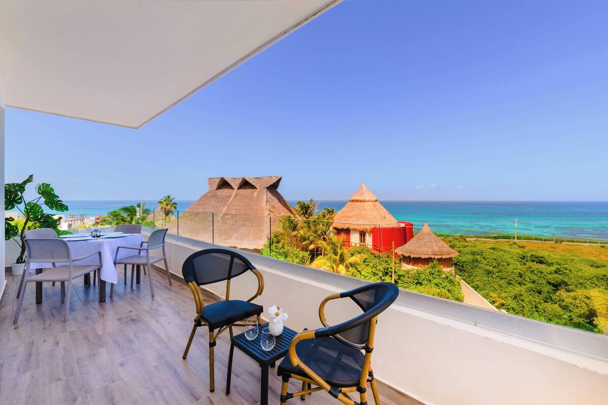 A balcony with a table and chairs overlooking a beach with thatched-roof huts and a clear blue sky.