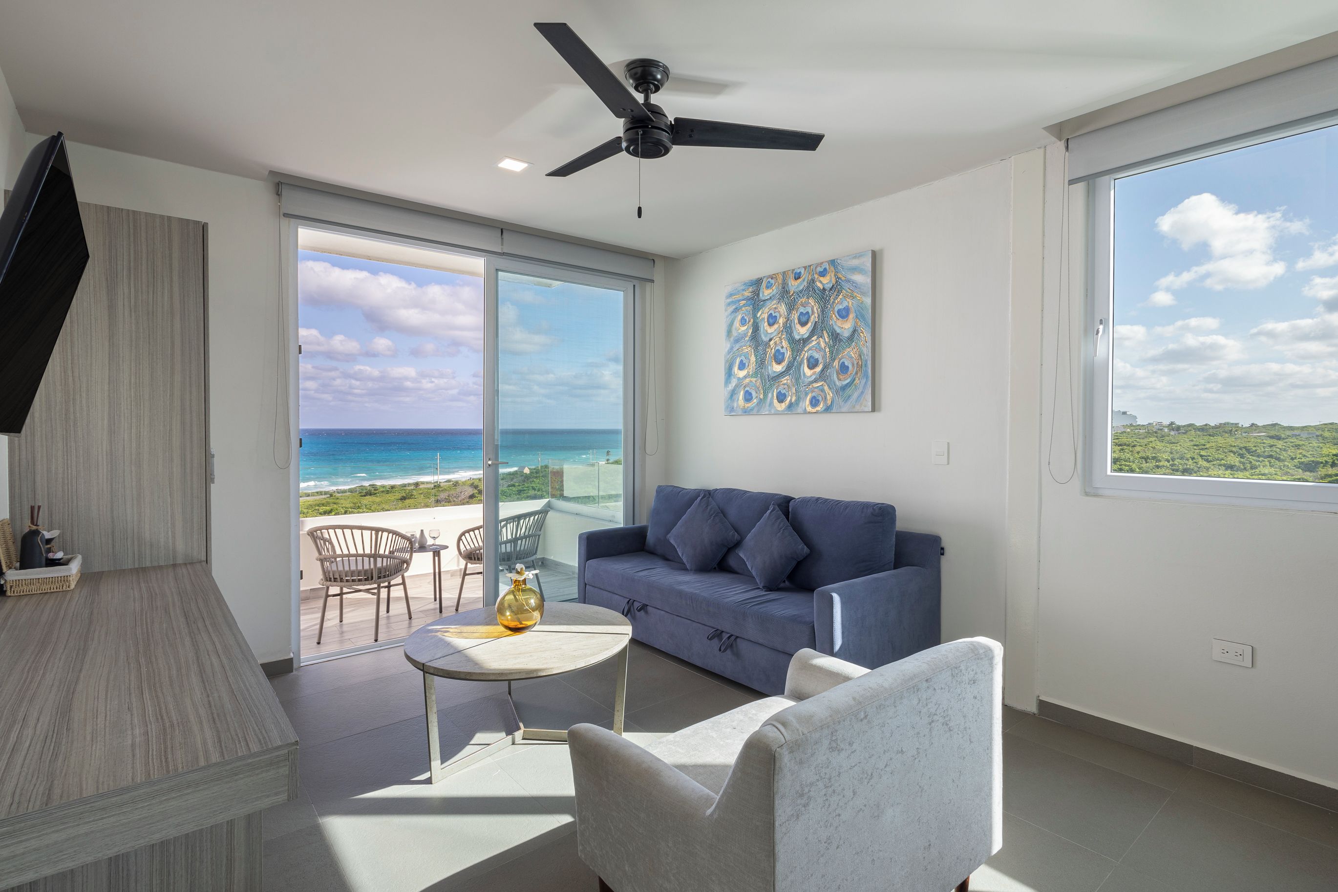 A modern living room with a large window overlooking a beach, a ceiling fan, and a blue sofa.