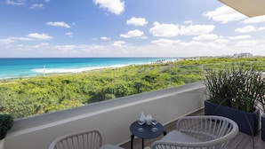 A balcony with wicker chairs and a small table, overlooking a beach and lush greenery.
