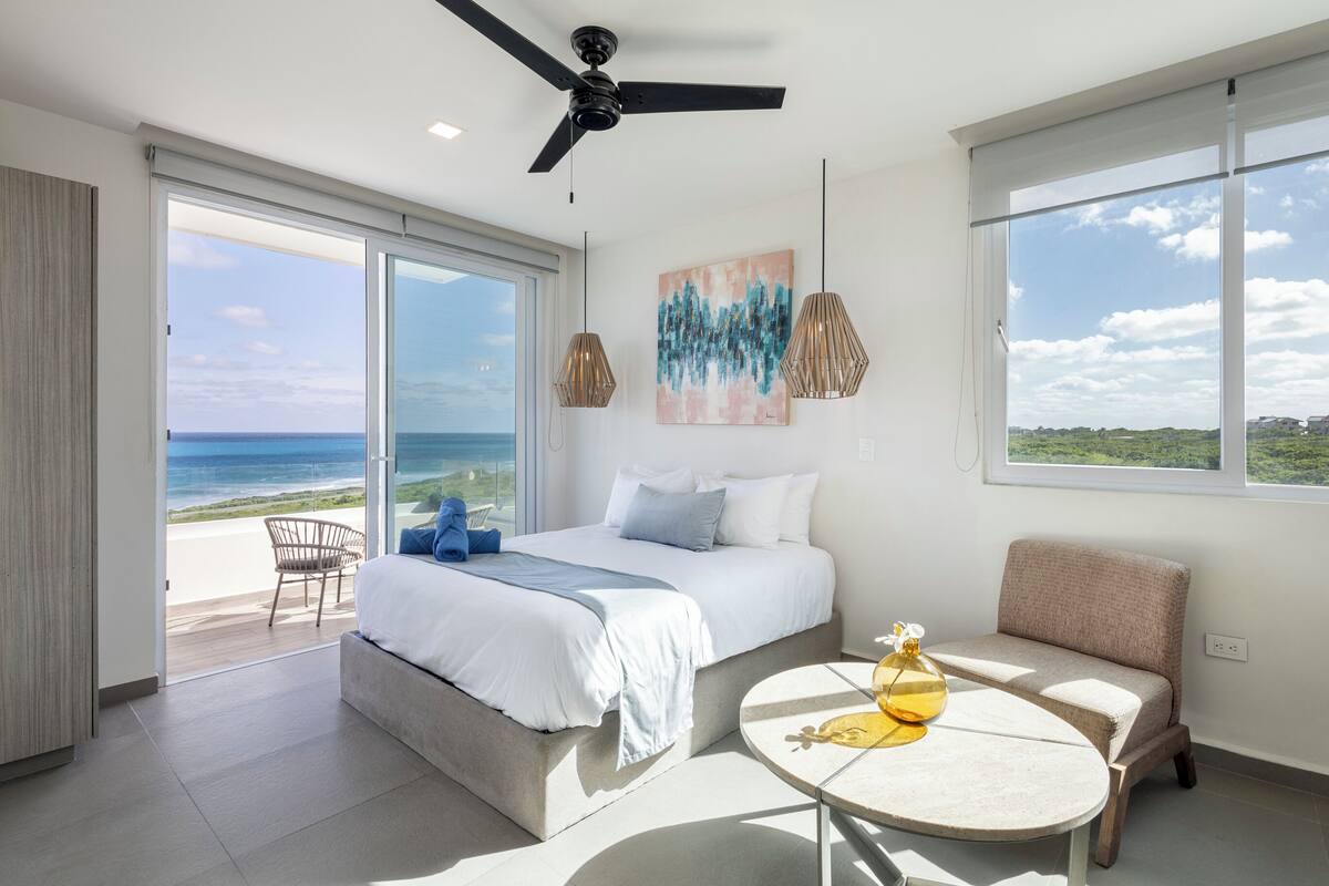 A modern bedroom with a large bed, a seating area, and a view of the ocean.