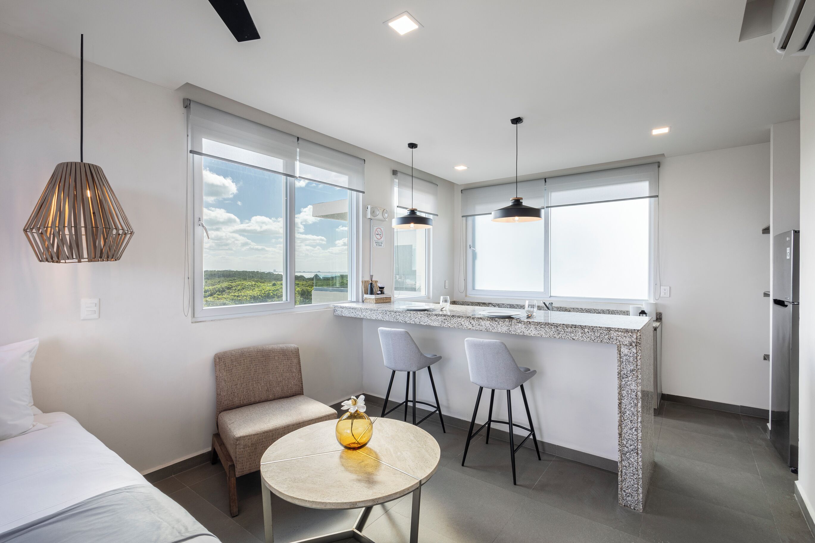 A modern kitchen with a large window, two bar stools, a round table with a vase, and a sofa.