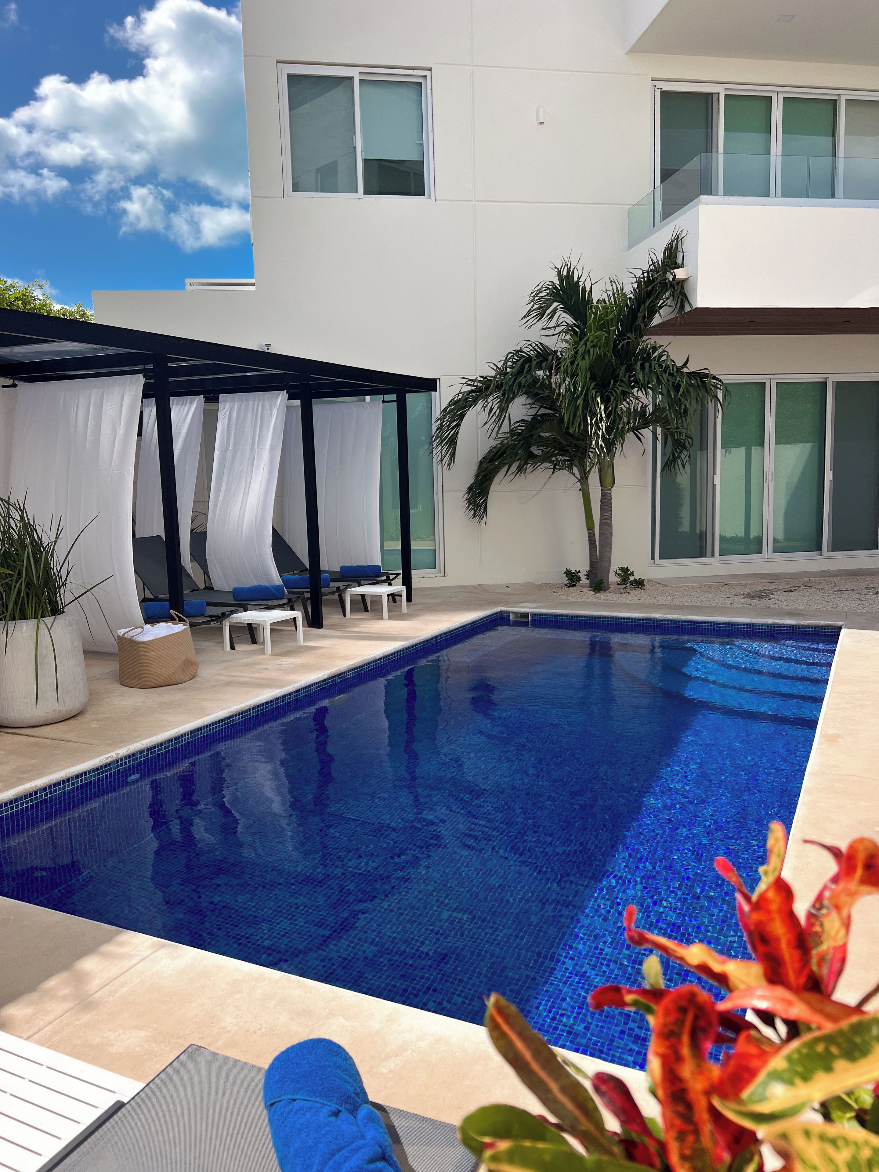 Un área de piscina moderna al aire libre con piscina azul cristalina, sillas de descanso y un espacio de estar cubierto.