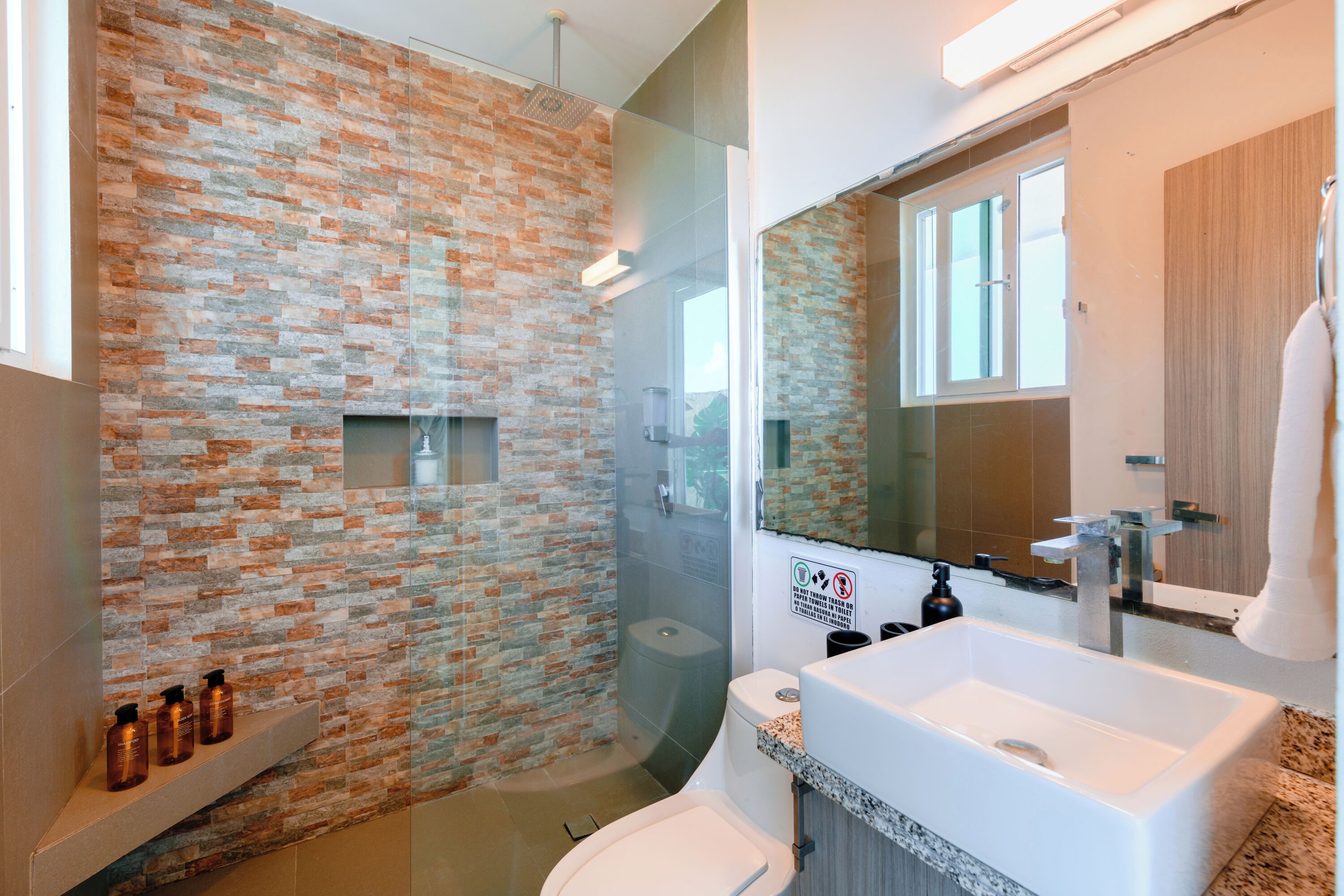 A bathroom with a brick wall, a glass shower enclosure, a white sink, and a mirror.