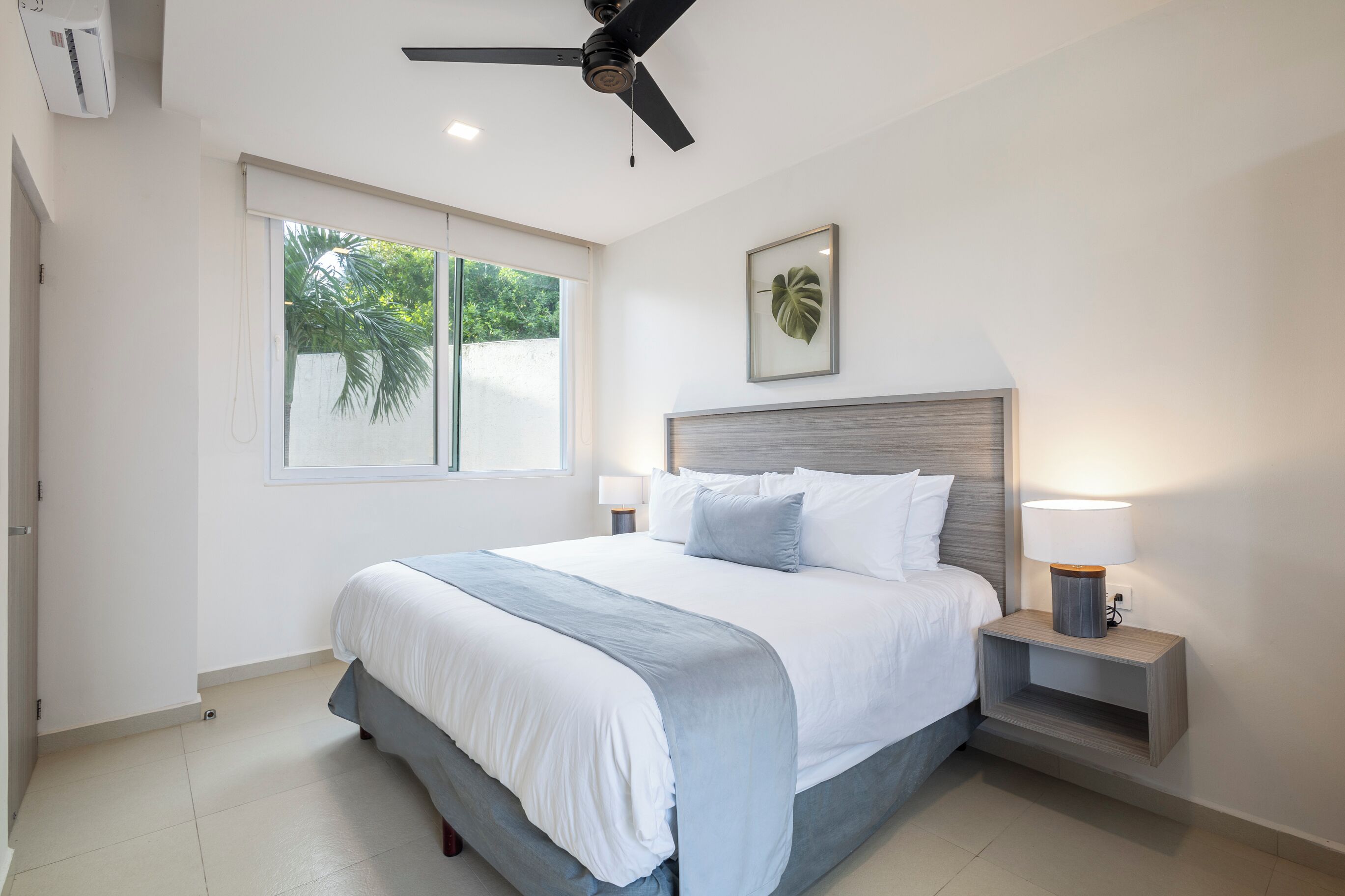 Une chambre moderne avec un lit, un ventilateur de plafond, une table de chevet avec une lampe, une fenêtre et une plante décorative dans un cadre.