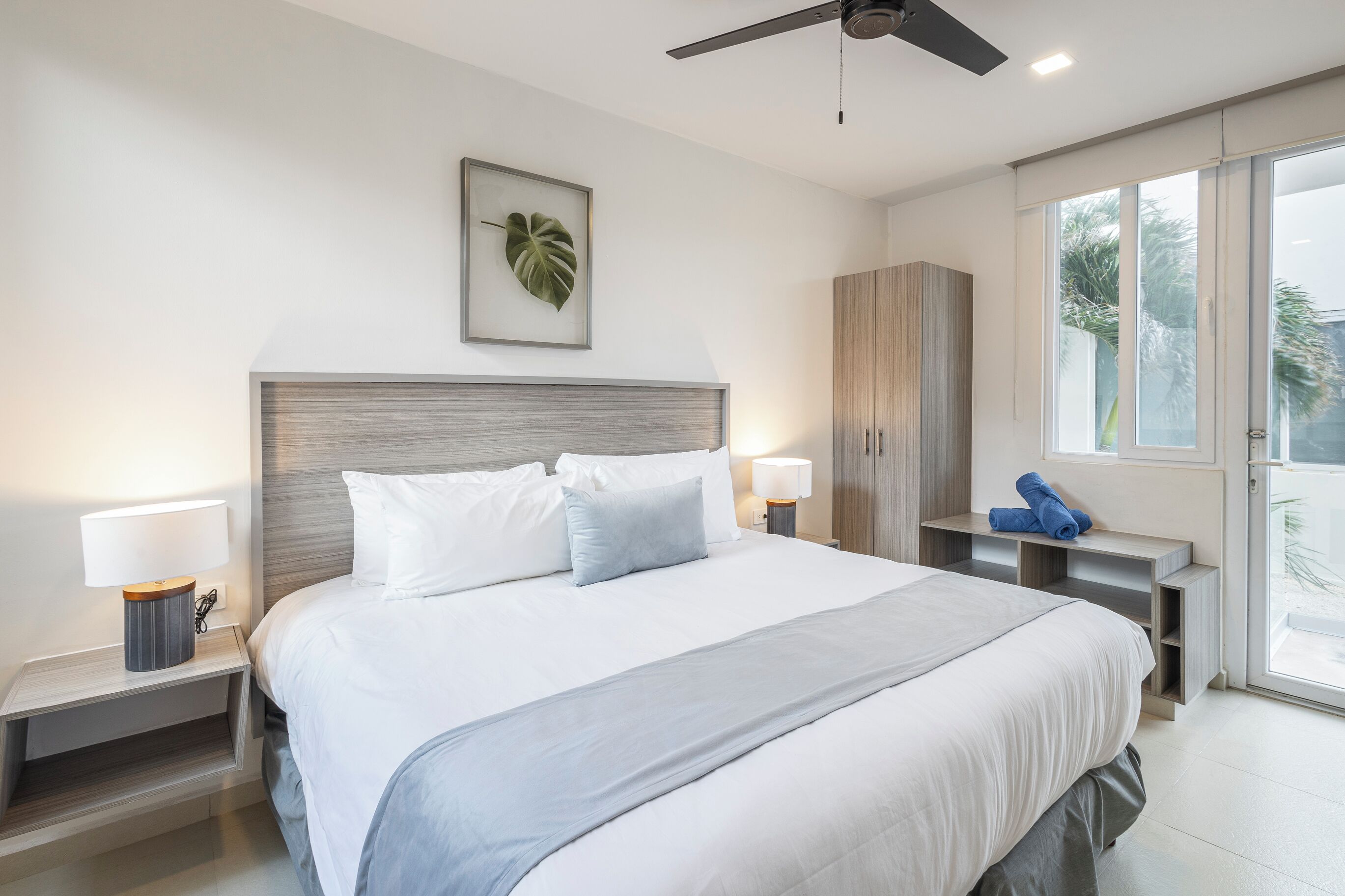 A modern hotel room with a large bed, two bedside tables with lamps, a ceiling fan, a wardrobe, and a view of the outdoors through a sliding door.