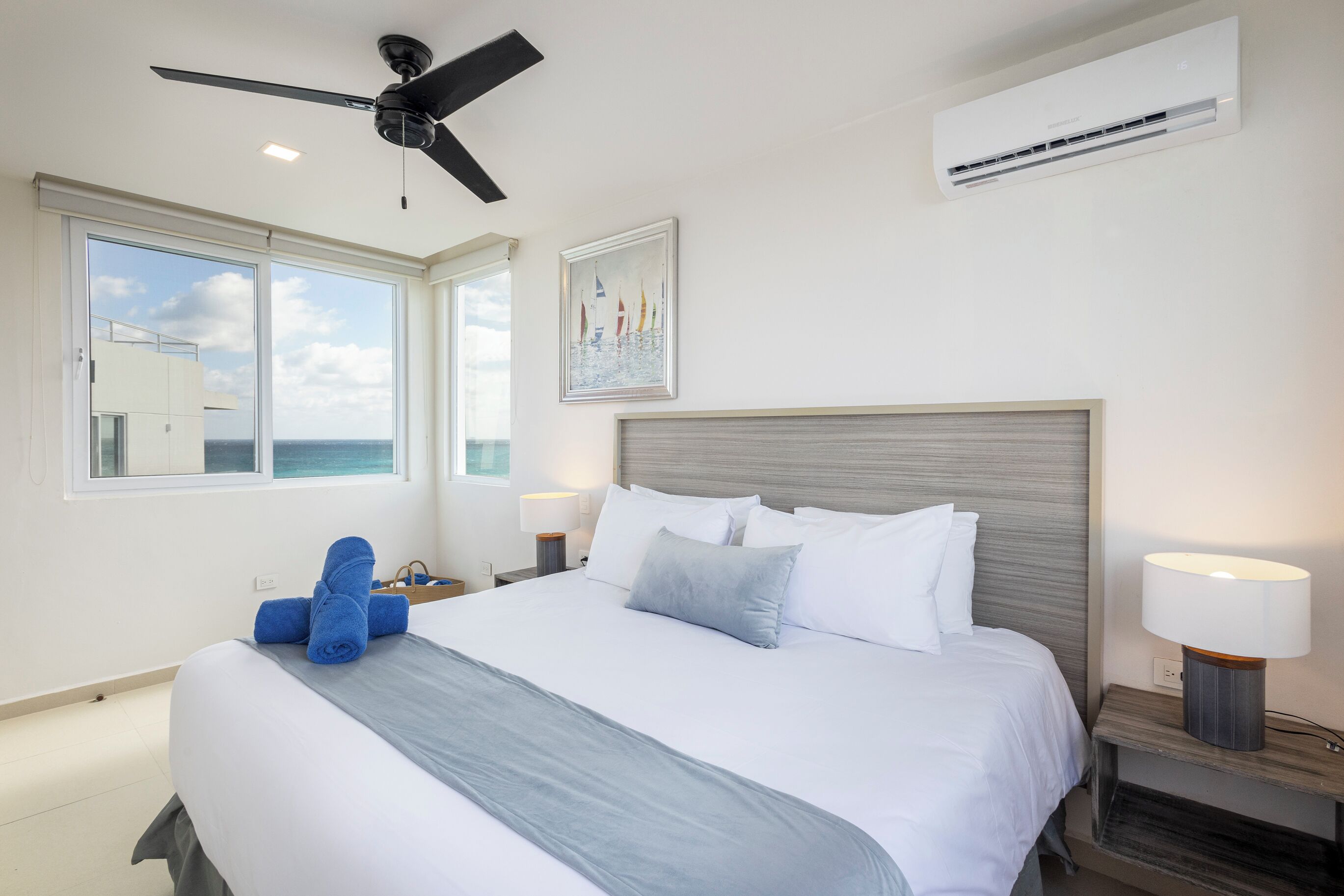 Ein Hotelzimmer mit einem großen Bett, einem Deckenventilator, einer Klimaanlage, einem Fenster mit Meerblick und einem Gemälde an der Wand.