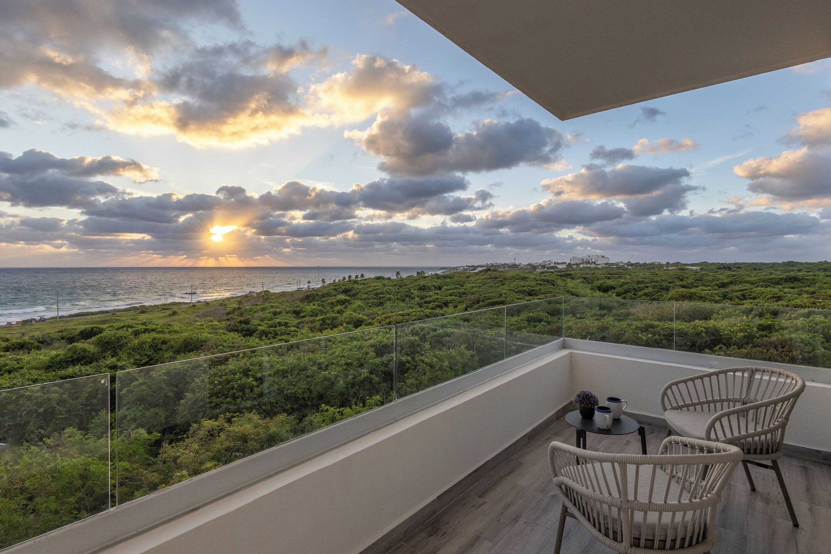 A balcony with wicker chairs, a small table, and a glass railing overlooking a coastal landscape with dense greenery and the ocean.