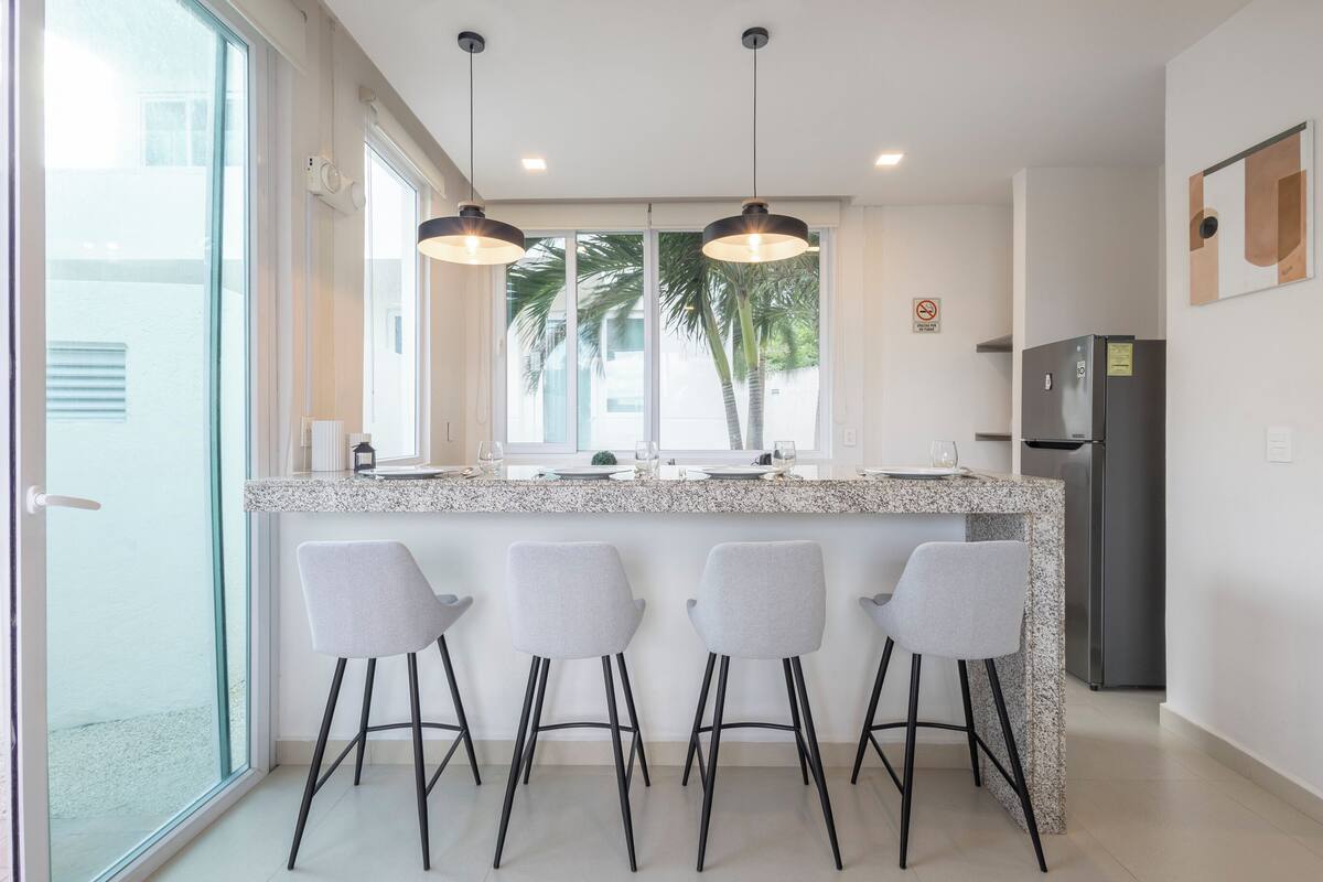A modern kitchen with a granite countertop, four bar stools, a refrigerator, and pendant lights.