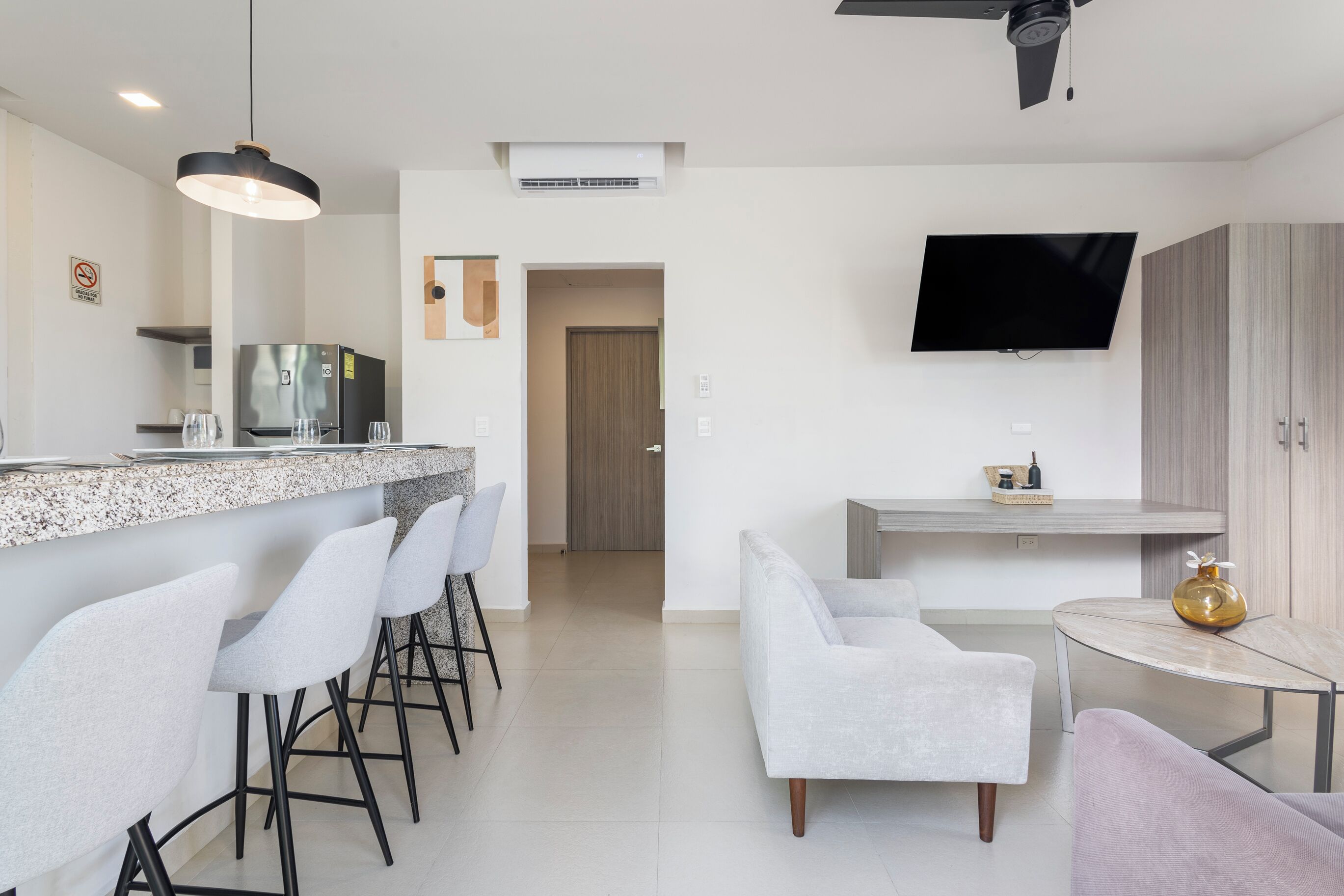 A modern kitchen with a granite countertop, bar stools, a built-in refrigerator, and a flat-screen TV mounted above a wooden cabinet.