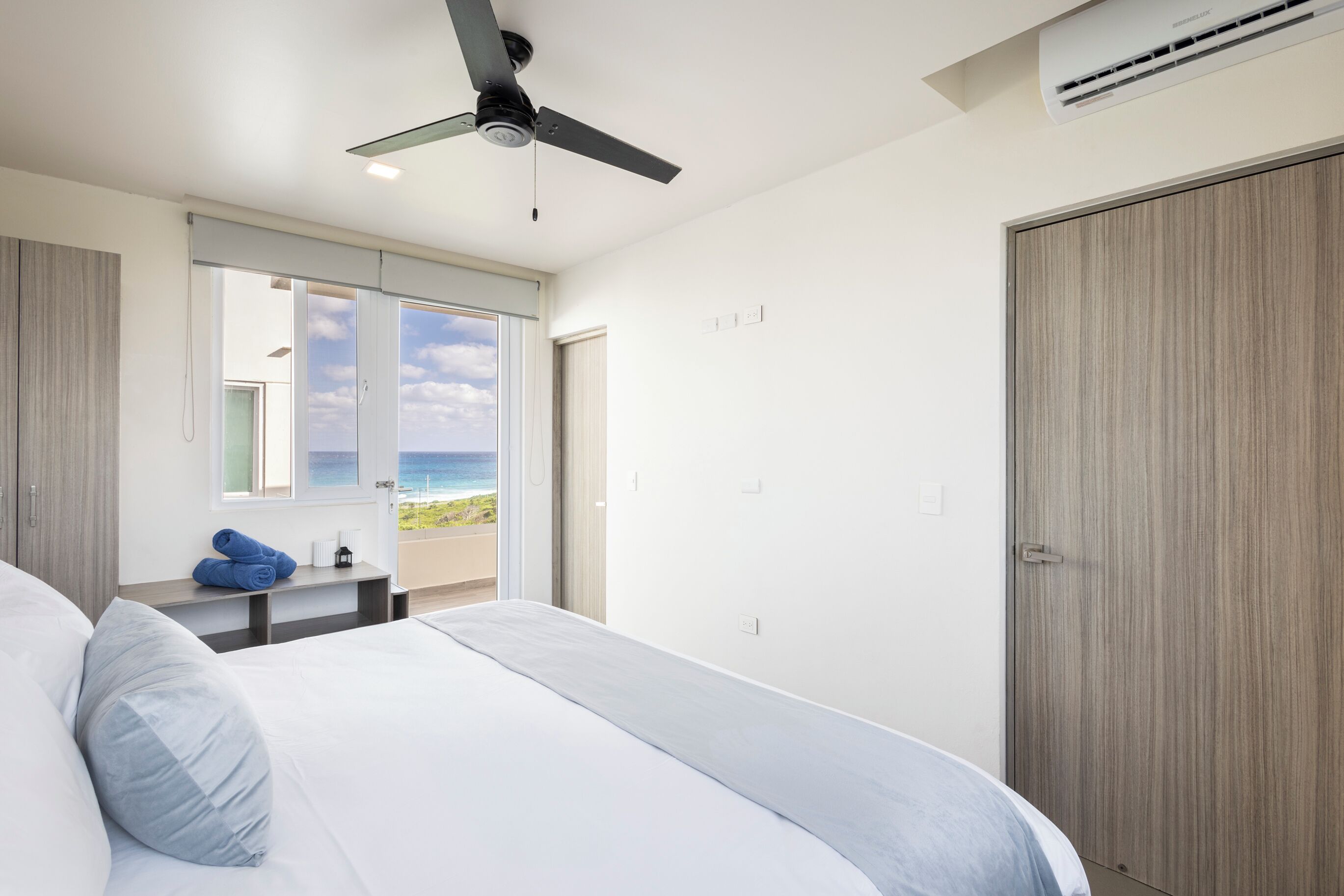A bedroom with a bed, a ceiling fan, an air conditioning unit, a window with a view of the beach, and a wooden wardrobe.