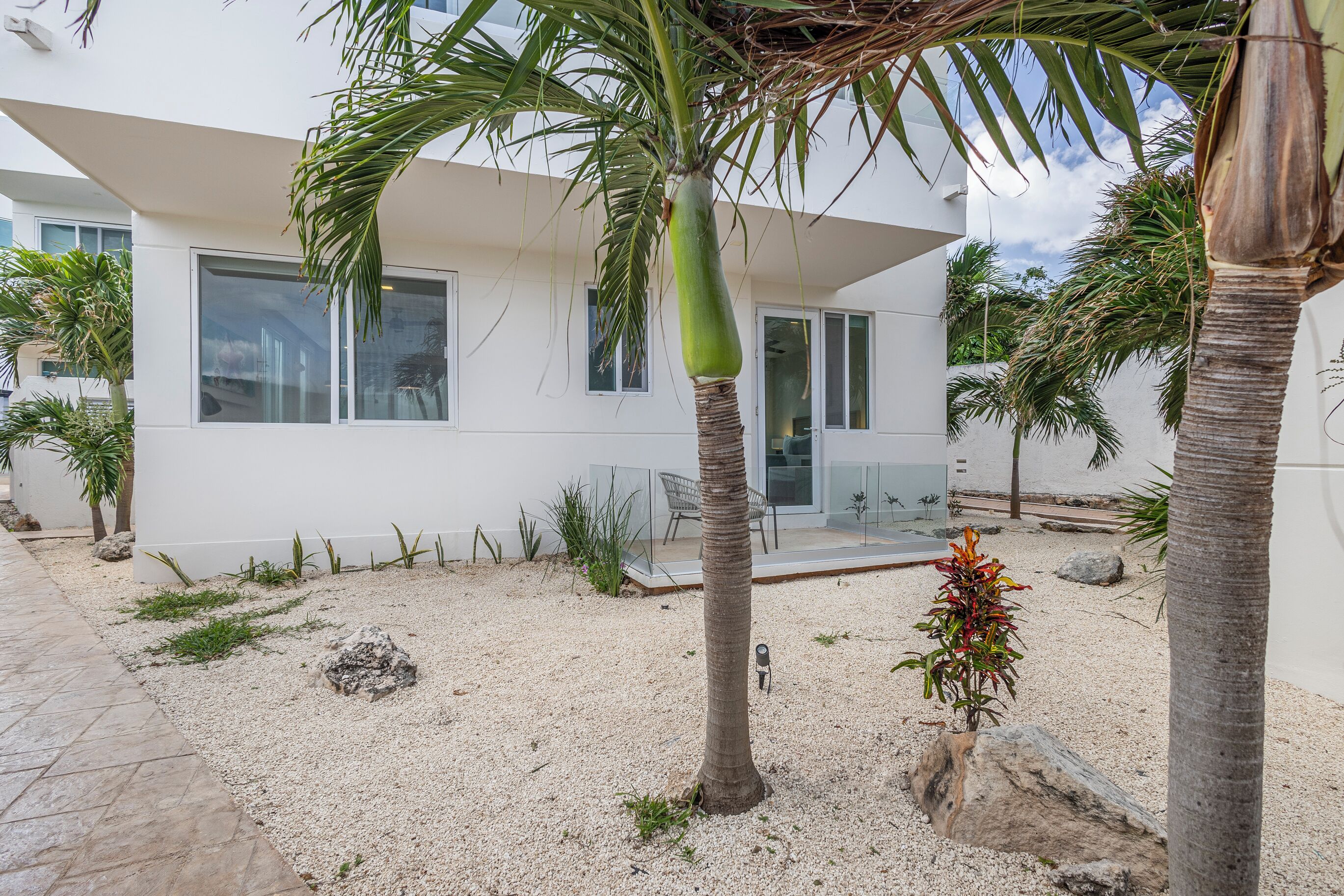 A modern house with a white exterior, large windows, and a sandy courtyard with palm trees and rocks.