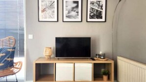 Un salon moderne avec un meuble TV en bois, trois tableaux encadrés accrochés au mur, un fauteuil en osier et une petite plante.