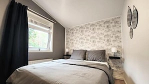 Une chambre avec un grand lit, une fenêtre avec des rideaux, une table de chevet avec une lampe et un mur recouvert d’un papier peint à motifs tropicaux.