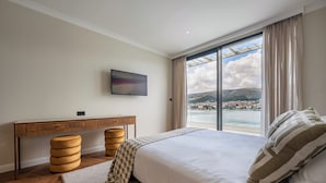 Ein Schlafzimmer mit einem großen Bett, einem Schreibtisch und Blick auf ein Gewässer und Hügel.