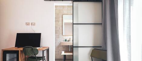 A modern hotel room with a desk, chair, TV, and a sliding door leading to a bathroom.