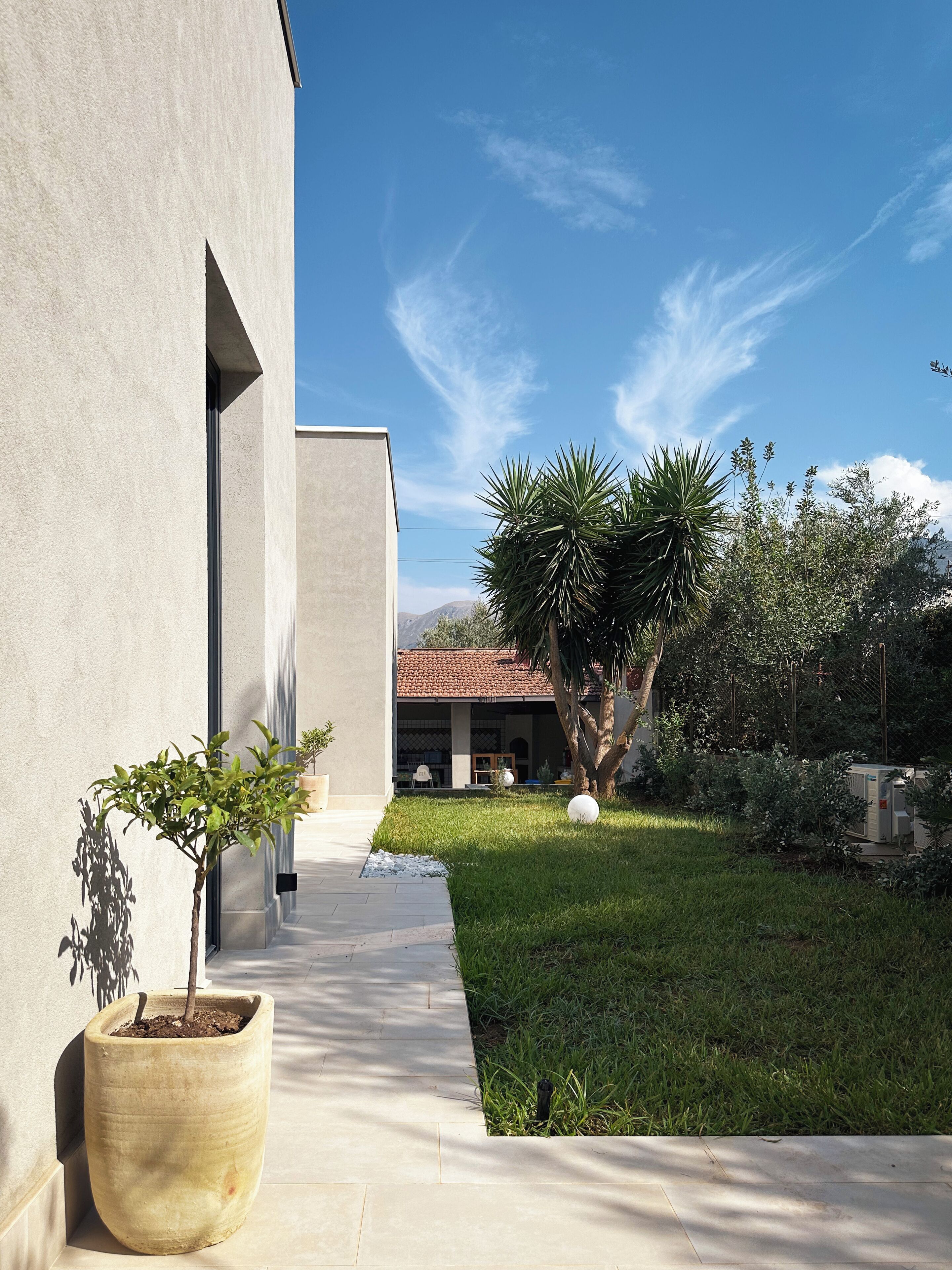 A modern building with a concrete facade, a paved pathway, and a well-maintained lawn with a palm tree and a potted plant.