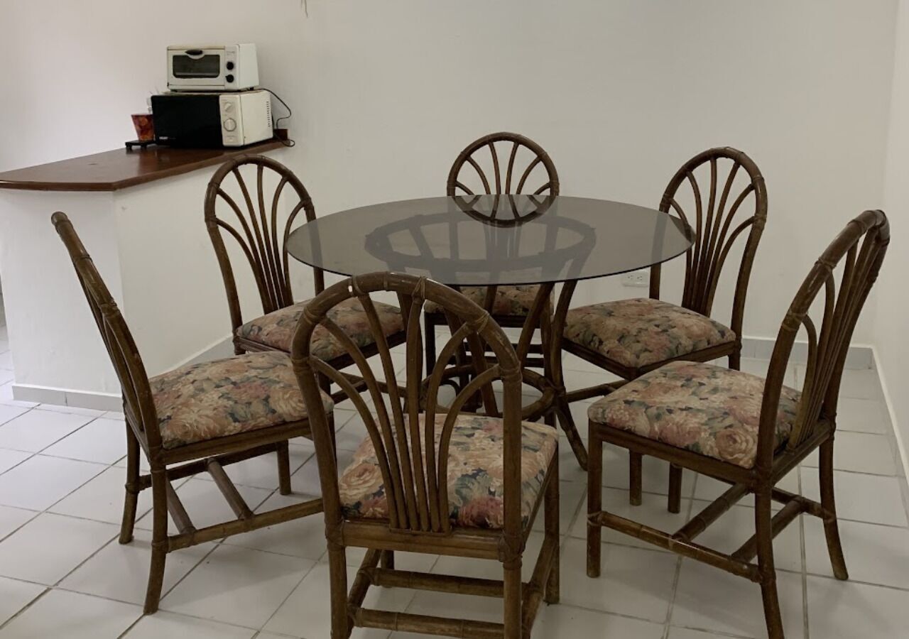 Une table ronde en verre, entourée de six chaises en bois dont les assises sont recouvertes d’un tissu à motifs floraux.