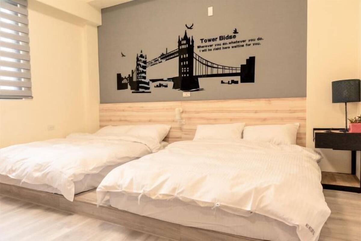 A hotel room with two beds, a wall mural of Tower Bridge, and a nightstand with a lamp.