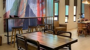 Un espace de salle à manger moderne avec une table et des chaises en bois, de grandes fenêtres équipées de stores et un mur décoré d’une œuvre abstraite aux couleurs vives.