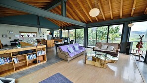 A spacious living area with a wooden ceiling, large windows, a sofa, a coffee table, and a bookshelf.