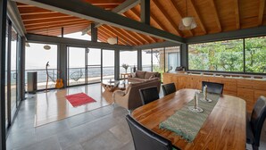 A spacious living area with a wooden ceiling, large windows, a wooden dining table, and a view of greenery outside.