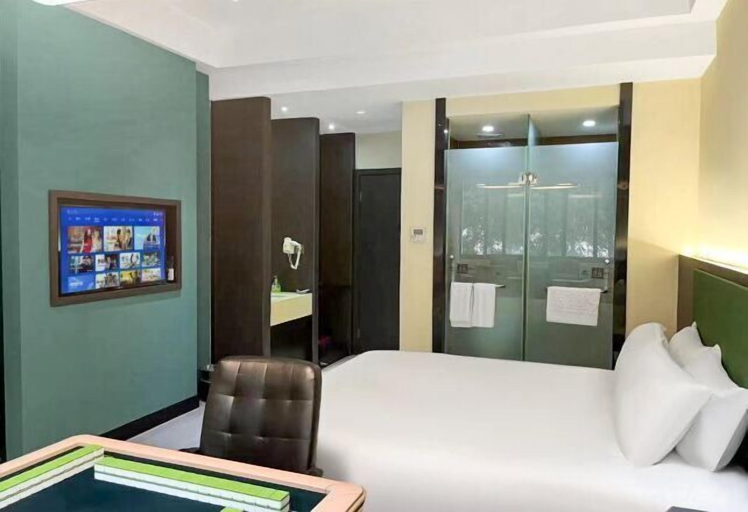 Une chambre d’hôtel comprenant un lit, un bureau, une chaise, une télévision et une salle de bain avec une douche en verre, ainsi qu’un mur vert.