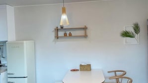 A kitchen with a white refrigerator, a hanging pendant light, a wooden shelf, and a dining table with chairs. - GLA Aewol (Jeju City)