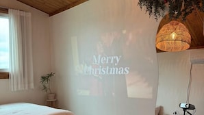 A bedroom with a bed, a projector screen displaying "Merry Christmas," a potted plant, and a hanging light fixture. - GLA Aewol (Jeju City)